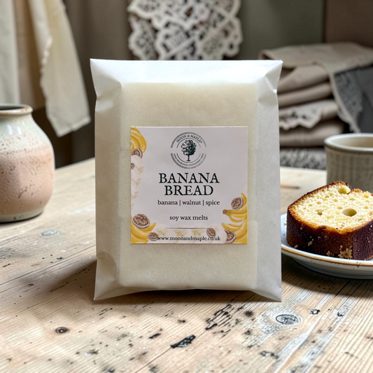 A package of Banana Bread soy wax melts displayed on a rustic wooden table next to a slice of banana bread, with cozy home decor in the background.