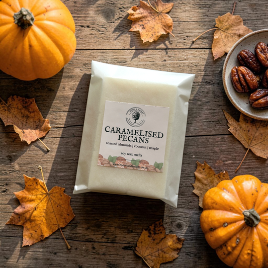 A package of caramelised pecans wax melts sits on a rustic wooden table surrounded by autumn leaves, small pumpkins, and a plate of pecans. 
