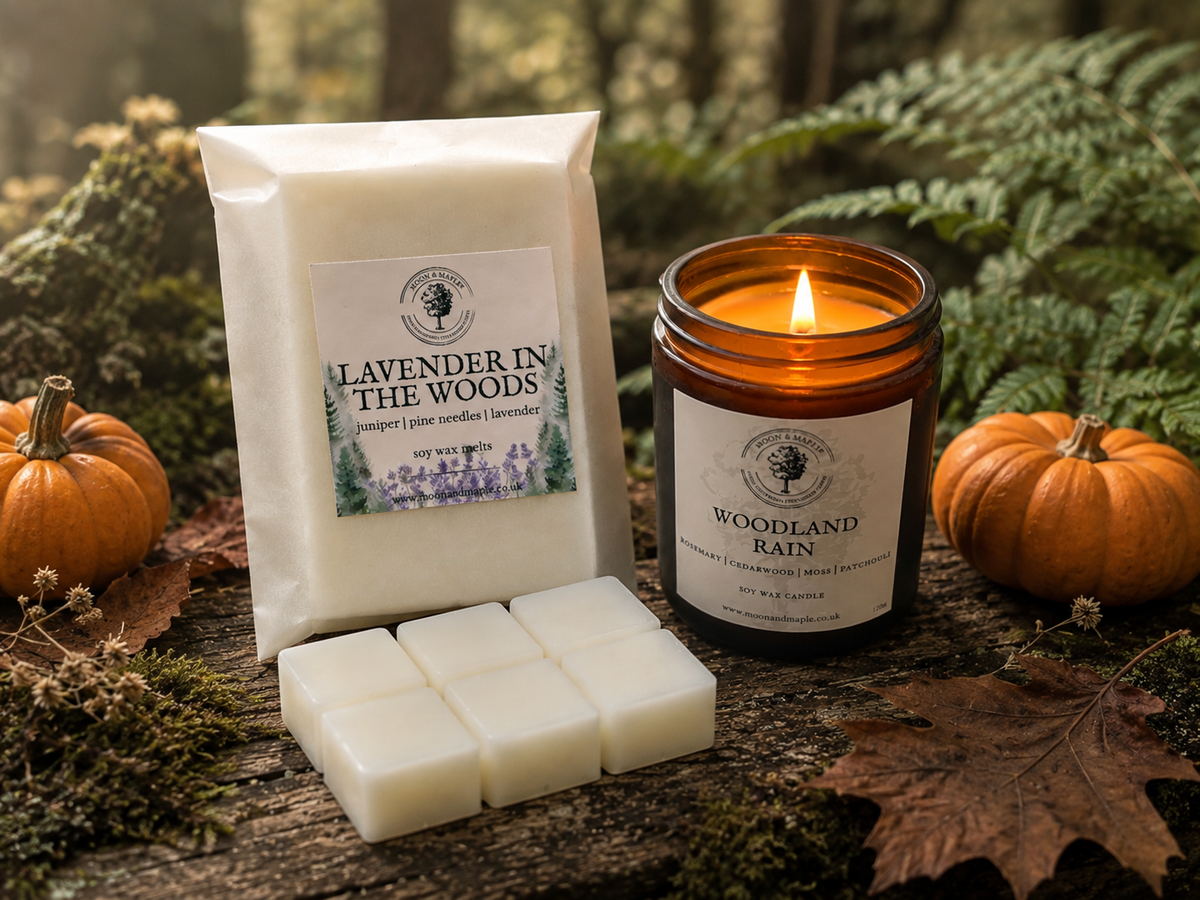 A candle and wax melt pack from the brand Moon & Maple, featuring a lit amber glass jar candle labeled 'Woodland Rain' and a white paper pack of 'Lavender in the Woods' wax melt cubes, styled on a rustic log with autumn leaves, moss, and small pumpkins.