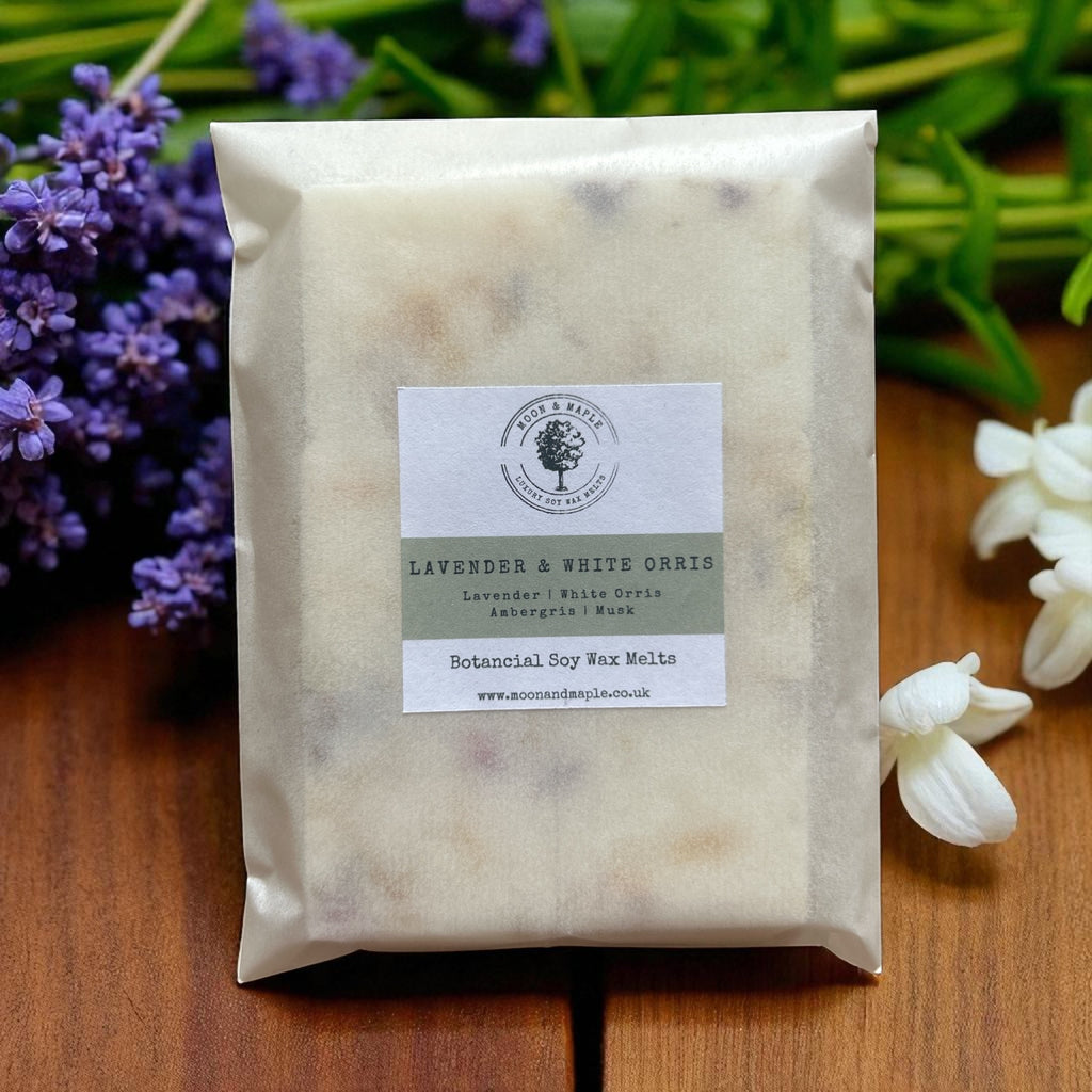 Packaged wax melts with a label on a wooden surface with lavender and white flowers in the background