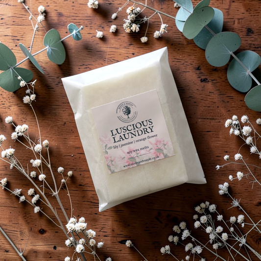 A flat-lay photograph of a white paper bag of Luscious Laundry wax melts on a rustic wooden table, decorated with eucalyptus leaves and white baby's breath flowers. The label on the front of the bag features a floral design with pink flowers and green leaves.
