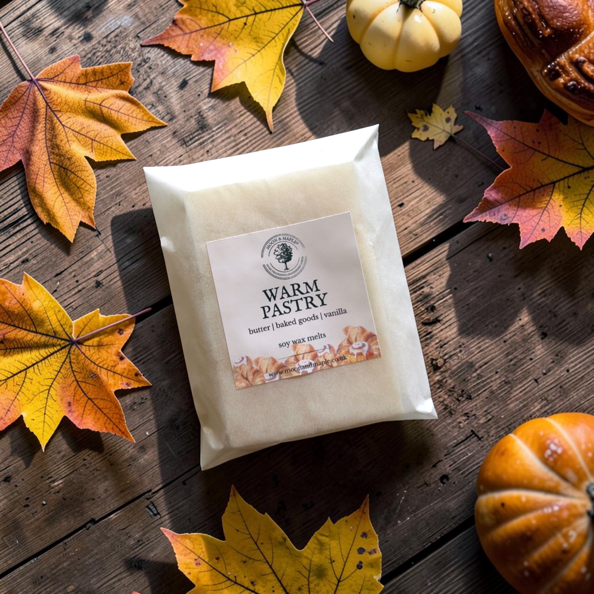 A package of Warm Pastry soy wax melts sits on a rustic wooden table surrounded by autumn leaves and small pumpkins.