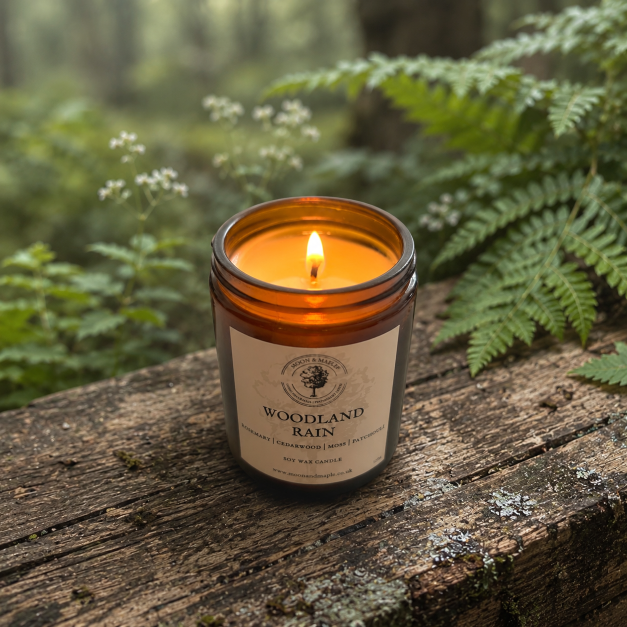 An amber glass jar candle with a lit flame sits on a weathered wooden surface outdoors, surrounded by green ferns and soft forest blur. The front label reads 'WOODLAND RAIN'